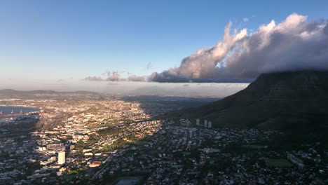 Amazing-aerial-shot-of-town-at-sunset-next-to-Table-mountain-with-clouds-hanging-overhead,-shadows-and-warm-light-creating-a-dramatic-look,-Cape-Town-South-Africa