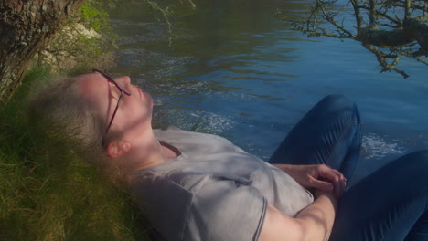 Woman-In-Glasses-Relaxing-With-Eyes-Closed-Lying-On-A-Grassy-Hillside-Next-To-A-Lake-On-A-Beautiful-Sunny-Day