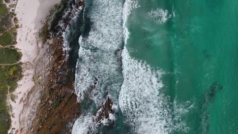 Turquoise-ocean-waves-wash-up-on-empty-rugged-Cape-Town-beach---aerial-top-down