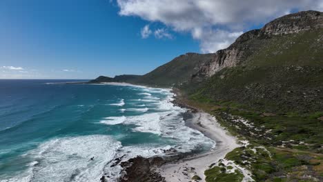 Aerial-dolly-shot,-of-waves-crashing-on-a-beach-below-rocky-cliffs-on-a-bright-sunny-day-with-partly-cloudy-sky,-Misty-Cliffs-near-Cape-Town-South-Africa