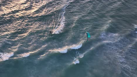 Aerial-birdseye-view-of-a-kite-surfer-at-sunset-sailing-over-cresting-white-waves-with-beautiful-sunlight-softly-reflected-off-the-water
