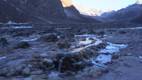 A-small-stream-in-a-canal-flowing-into-a-pipe-surrounded-by-frosty-grass-with-mountains-in-the-background