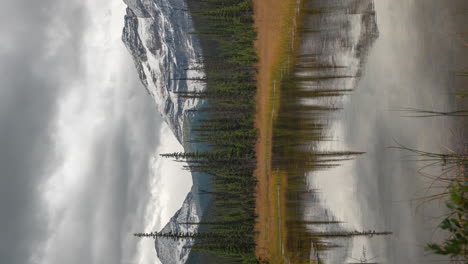 Vertical-Time-Lapse,-Cold-Autumn-Day-in-Mountains-of-Canada,-Dramatic-Dark-Clouds-Above-Snow-Capped-Peaks-and-Valley-With-Conifer-Forest-and-Lake-Water