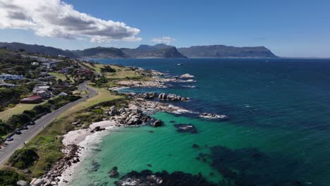 Beautiful-aerial-dolly-shot-of-a-road-along-the-rocky-coast-with-deep-green-waters,-houses,-mountains-and-cliffs-in-the-background-on-a-sunny-day-near-Cape-Town-South-Africa