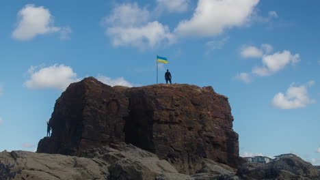 People-Climb-and-Stand-on-Chapel-Rock-with-Ukrainian-Flag-on-a-Sunny-Day,-Perranporth,-Cornwall,-England,-UK---timelapse