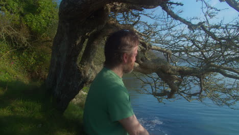 Bearded-Mid-Adult-Man-Enjoys-Warm-Morning-Sunshine-Sitting-Beside-Tree-Looking-into-The-Lake