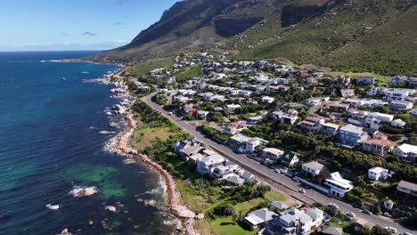 Aerial-orbit-of-vacation-homes-on-the-coast-of-a-scenic-ocean-vista-with-steep-mountains,-rocky-shore-and-sea-green-waters-near-Cape-Town-South-Africa