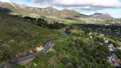 Reverse-aerial-dolly-shot-of-cars-and-buses-driving-on-a-curvy-mountain-road-above-a-town-with-clouds-and-shadows,-near-Cape-Town-South-Africa
