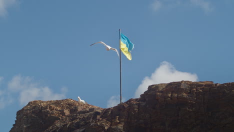 Möwen,-Die-Vom-Kapellenfelsen-Wegfliegen,-Mit-Ukrainischer-Flagge,-Die-In-Der-Meeresbrise-Weht---Statisch