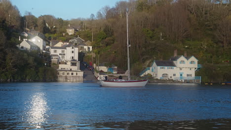 Sailboat-Cruising-through-Glistening-Fowey-River,-Cornwall-with-Historic-Daphne-du-Maurier-house-in-background---wide-shot