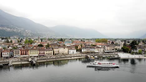 Aerial-flyover-alongside-the-waterfront-of-Ascona-in-Ticino,-Switzerland-with-a-view-of-a-boat-arriving-at-the-pier-and-the-city-buildings-along-the-lakeside-promenade-on-the-shores-of-Lago-Maggiore