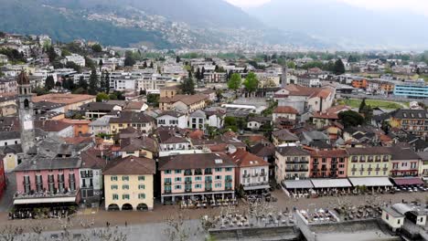 Aerial-flyover-over-the-rooftops-of-Ascona-in-Ticino,-Switzerland-towards-the-shores-of-Lago-Maggiore,-with-a-boat-departing-from-the-waterfront-towards-the-lake-with-a-view-of-the-Alps