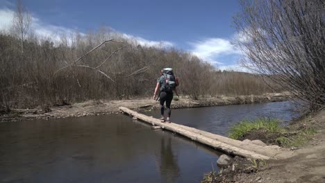 Female-hiker-with-backpacking-crossing-small-river-on-log,-New-Mexico-USA