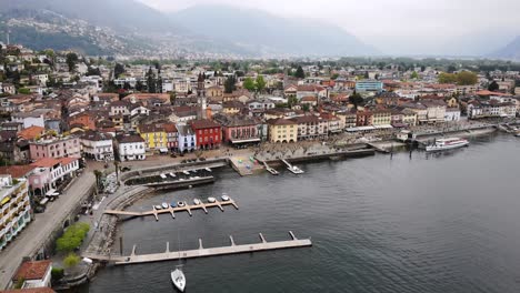Aerial-flyover-over-the-shores-of-Lago-Maggiore-towards-the-rooftops-of-Ascona-in-Ticino,-Switzerland-with-a-view-of-the-lakeside-promenade,-boats-and-church-tower