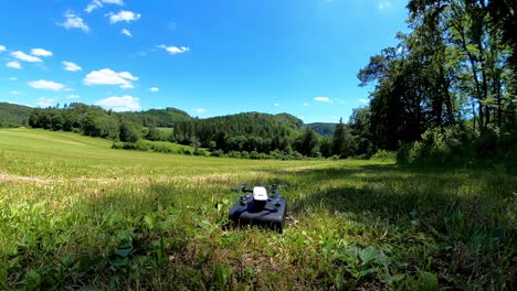 Kleine-Weiße-Drohne-Mit-Kamera-Hebt-Aus-Schwarzer-Tasche-Ab,-Sonnige-Tageslandschaft-Im-Naturhintergrund-Mit-Wald-Und-Grasbewachsener-Wiese