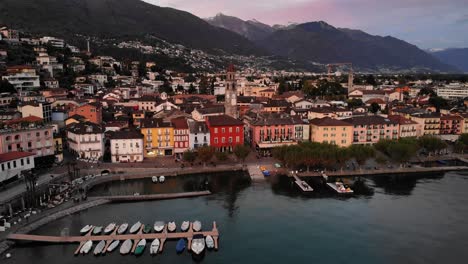 Aerial-flyover-over-the-shores-of-Lago-Maggiore-towards-the-promenade-and-church-tower-of-Ascona,-Switzerland-with-a-view-of-mountains,-boats,-colorful-houses