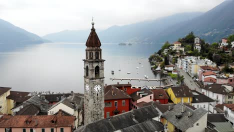 Aerial-flyover-around-the-belfry-tower-of-the-Chiesa-dei-Santi-Pietro-e-Paolo-church-and-over-the-rooftops-Ascona,-Switzerland-revealing-the-waters-of-Lago-Maggiore-and-Alps-in-the-background