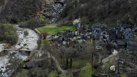 Aerial-flyover-over-Foroglio-in-Ticino,-Switzerland-with-a-pan-up-motion-up-from-the-rooftops-of-the-old-stone-houses-in-the-village-up-to-the-waterfall-at-dusk