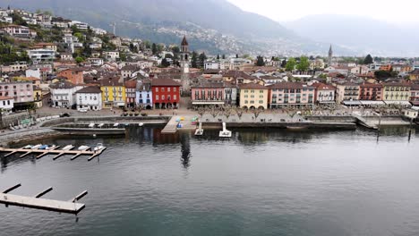 Aerial-flyover-along-the-shore-of-Lago-Maggiore-and-promenade-of-Ascona-in-Ticino,-Switzerland-with-a-turn-over-the-city-rooftops-revealing-the-lake-and-mountains-behind-the-church-tower
