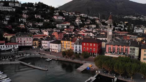 Aerial-flyover-along-the-lakeside-promenade-of-Ascona,-Ticino-on-the-shores-of-Lago-Maggiore-in-Italian-Switzerland-at-the-end-of-a-summer-day-with-colorful-houses,-church-tower-and-boat-pier-in-view