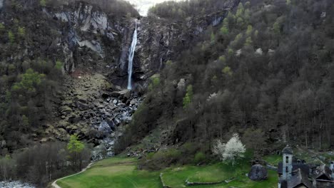 Aerial-flyover-with-a-side-of-the-old-stone-house-of-Foroglio-village-in-Ticino,-Switzerland-underneath-the-waterfall-at-dusk