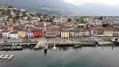 Aerial-flyover-with-a-turn-from-the-shores-of-Lago-Maggiore-over-the-promenade-of-Ascona-in-Ticino,-Switzerland-over-the-rooftops-around-the-church-tower-revealing-the-lake-and-mountains
