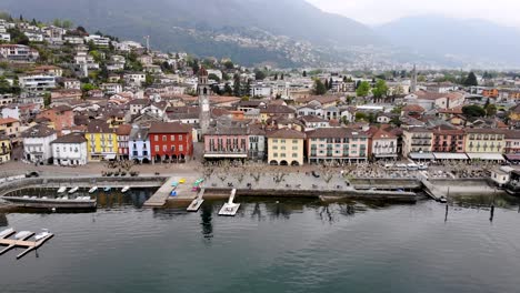 Aerial-flyover-with-a-turn-from-the-shores-of-Lago-Maggiore-over-the-promenade-of-Ascona-in-Ticino,-Switzerland-over-the-rooftops-around-the-church-tower-revealing-the-lake-and-mountains