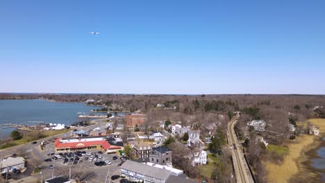 Plane-flies-across-a-blue,-clear-sky-without-clouds-over-a-small-town-next-to-a-lake-with-train-station