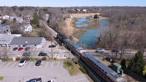 Train-on-its-tracks-through-small-village-diagonally-next-to-marsh-with-stopped-cross-traffic,-static-drone-shot