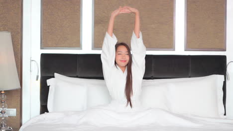 Pretty-asian-woman-waking-up-in-bed,-raising-and-stretching-arms-on-bright-morning,-slow-motion