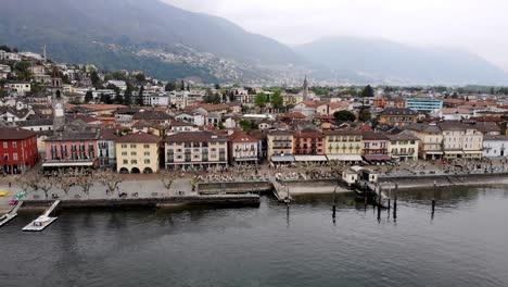 Aerial-flyover-along-the-promenade-of-the-city-of-Ascona-in-Ticino,-Switzerland-at-the-shores-of-Laggo-Maggiore-with-a-side-view-of-colorful-houses,-rooftops,-and-boats-on-a-cloudy-spring-day