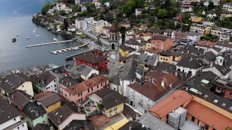 Aerial-flyover-over-the-rooftops-of-Ascona,-Switzerland-with-a-view-of-the-church-tower-and-shores-of-Lago-Maggiore-filled-with-boats-and-corwds-on-the-promenade