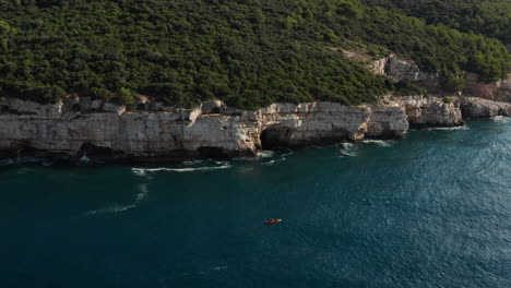 Aerial-Pullback-Shot-Of-A-Kayak-Sailing-Beside-A-Cliff-And-Dense-Greenery-Near-Pula-Istria-In-Croatia