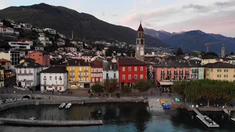 Aerial-flyover-along-the-lakeside-promenade-of-Ascona,-Ticino-on-the-shores-of-Lago-Maggiore-in-Italian-Switzerland-at-the-end-of-a-summer-day-with-colorful-houses-and-church-tower-in-view