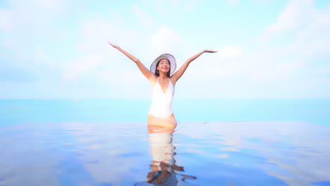 Happy-Exotic-Woman-in-Infinity-Swimming-Pool-Merging-With-Sea-and-Sky,-Raising-Arms-Full-of-Happiness