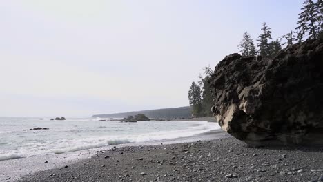 Slow-motion-of-sea-waves-hitting-rugged-sand-shore-near-woodland-on-an-overcast-day,-Sombrio-Beach,-British-Columbia,-Canada