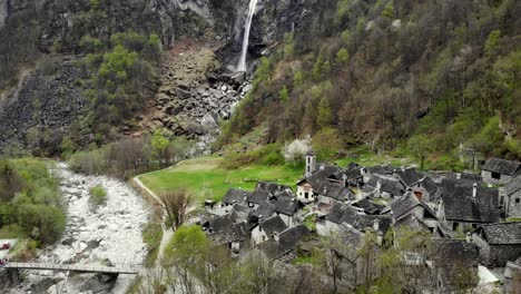 Aerial-flyover-over-the-rooftops-of-the-old-stone-houses-in-the-village-of-Foroglio-towards-the-waterfall-in-the-spring-time-alpine-landscape