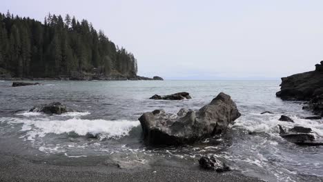 Slow-motion-of-foamy-sea-waves-hitting-rocky-shore,-woodland-in-background-on-an-overcast-day,-Sombrio-Beach,-British-Columbia,-Canada