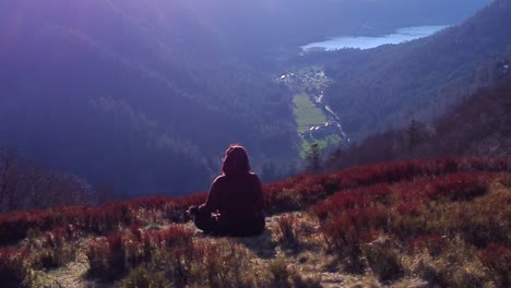 Female-meditating-in-lotus-position-at-the-top-of-a-mountain-at-sunset-relaxing-orbiting-view-over-lake-with-flare