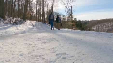 Two-young-hikers-with-backpacks-walking-on-snowy-road-during-winter