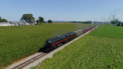 An-Aerial-View-Traveling-In-Front-of-An-Antique-Steam-Passenger-Train,-Blowing-Smoke-Traveling-Thru-Corn-Fields-and-Rich-Farmlands-on-a-Cloudless-Summer-Day