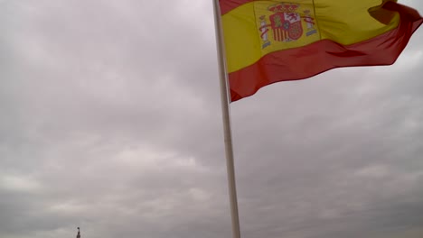Slow-tilt-down-from-Spanish-flag-waving-against-gray-sky-with-clouds-and-city-in-distance