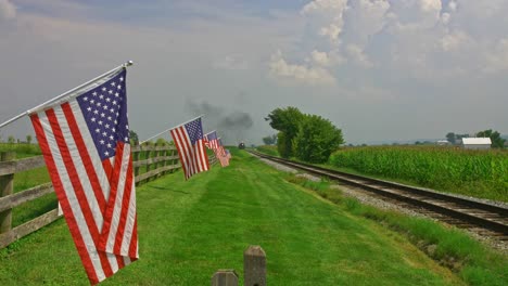 A-View-of-a-Line-of-Gently-Waving-American-Flag-on-a-Fence-by-Farmlands-as-a-Steam-Passenger-Train-Blowing-Smoke-Approaches-in-Late-Afternoon