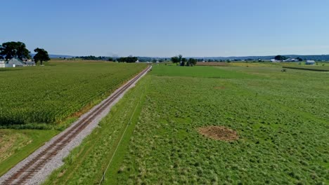 Eine-Luftaufnahme-Von-Fruchtbarem-Ackerland-Und-Maisfeldern-Entlang-Einer-Einzigen-Eisenbahnstrecke-An-Einem-Wolkenlosen-Sommertag
