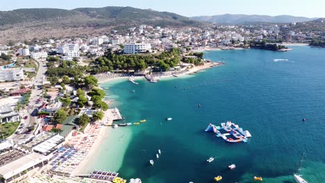 Ksamil,-Albania---Reversing-Aerial-of-Popular-Holiday-Destination-with-Hotels,-Beachclub,-Sunbeds,-Water-Playground-and-Island