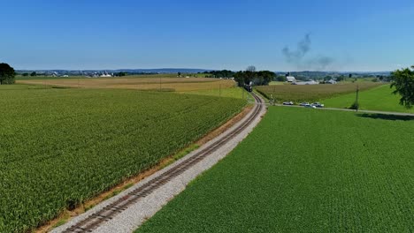 An-Aerial-View-Traveling-In-Front-of-An-Antique-Steam-Passenger-Train,-Blowing-Black-Smoke-Traveling-Thru-Corn-Fields-and-Rich-Farmlands-on-a-Cloudless-Summer-Day