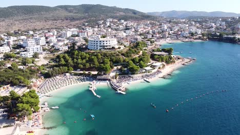 Ksamil,-Albania---Antena-De-Marcha-Atrás-De-Un-Popular-Destino-De-Vacaciones-Con-Hoteles,-Tumbonas,-Playa,-Mar-Y-Juegos-Acuáticos