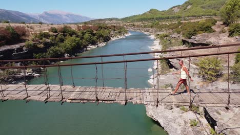 Tourist-Man-Walks-across-Wooden-and-Wobbly-Suspension-Bridge-at-Vjosa-River,-Albania---Aerial-Trucking-Sideways