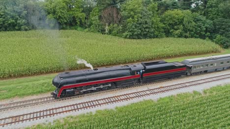 An-Aerial-Front-to-Side-View-of-a-Steam-Passenger-Train-Blowing-Smoke-as-it-Starts-to-Pull-out-of-a-Small-Station-Waiting-For-Passengers-to-Board-on-a-Summer-Day