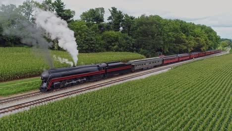An-Aerial-Front-to-Side-View-of-a-Steam-Passenger-Train-Blowing-Smoke-at-a-Small-Station-Waiting-For-Passengers-to-Board-on-a-Summer-Day